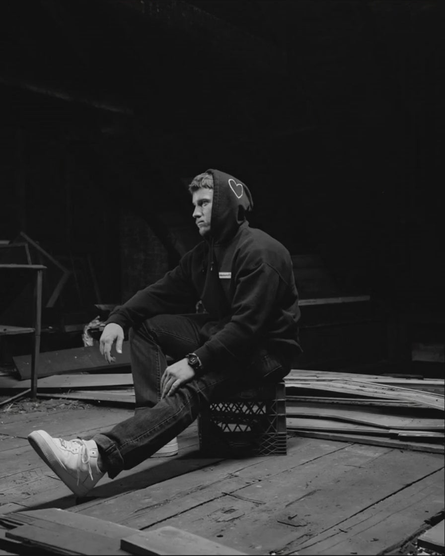 Ryan Zakrzewski seated in profile in a stripped-out attic, hood up, one leg extended